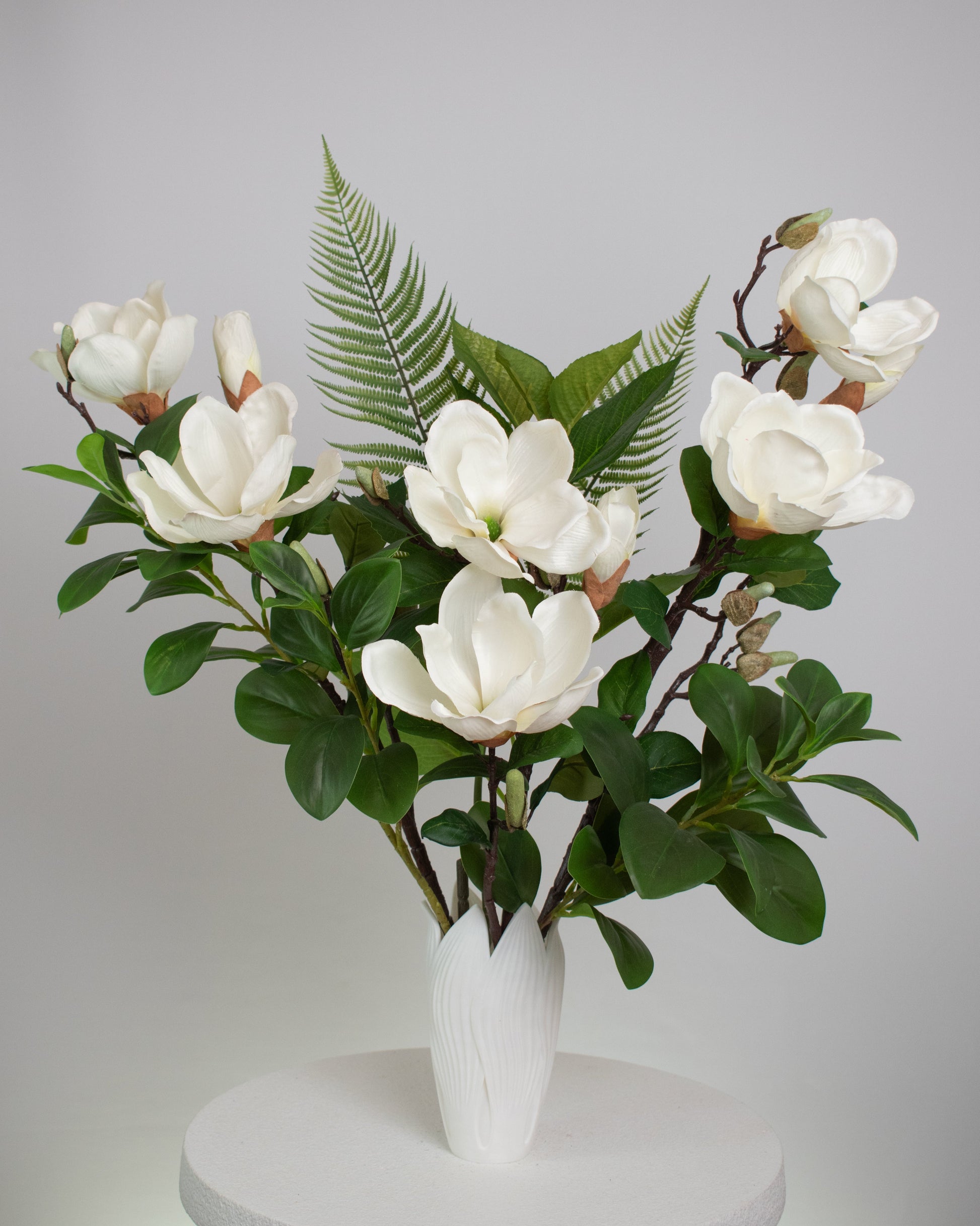 Artificial white magnolia flower arrangement with lush green leaves and fern accents, beautifully styled in a sleek white vase on a round white table, set against a neutral background.