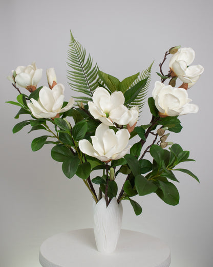 Artificial white magnolia flower arrangement with lush green leaves and fern accents, beautifully styled in a sleek white vase on a round white table, set against a neutral background.