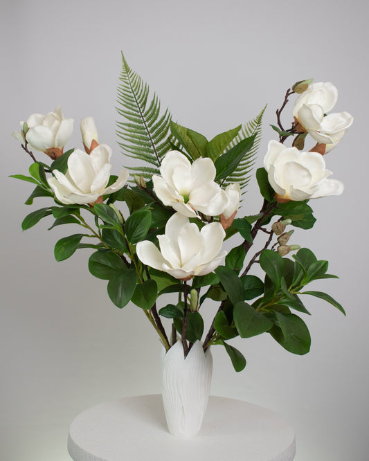 Artificial white magnolia flower arrangement with lush green leaves and fern accents, beautifully styled in a sleek white vase on a round white table, set against a neutral background.