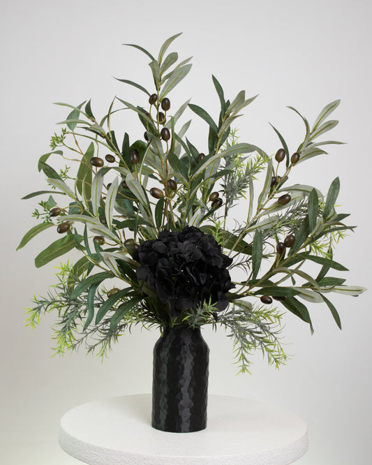 Artificial floral arrangement featuring lifelike olive branches with dark berries and a black hydrangea bloom, elegantly displayed in a textured black vase on a marble-topped round table against a neutral background.