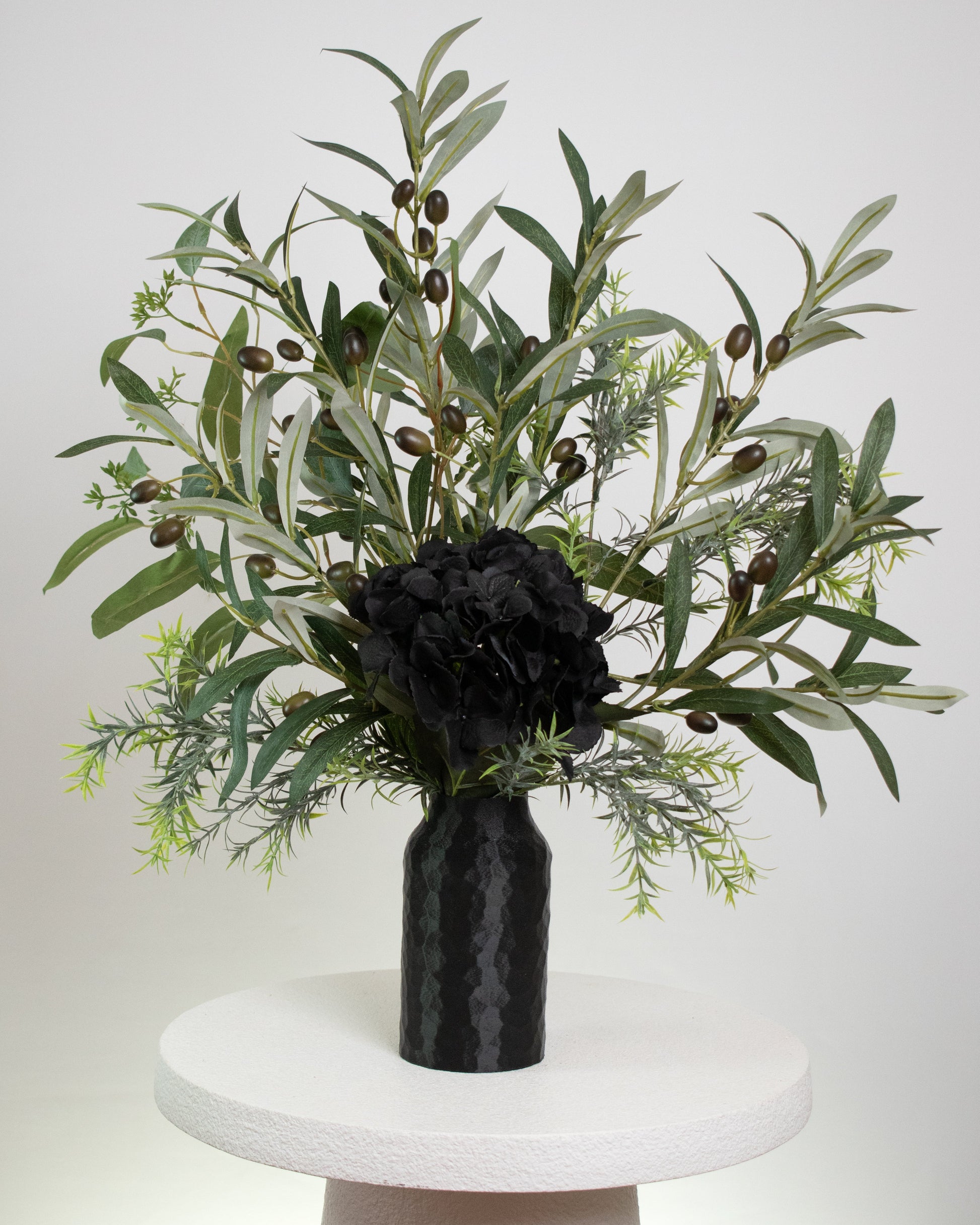 Artificial floral arrangement featuring lifelike olive branches with dark berries and a black hydrangea bloom, elegantly displayed in a textured black vase on a marble-topped round table against a neutral background.