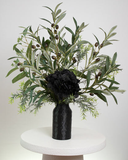 Artificial floral arrangement featuring lifelike olive branches with dark berries and a black hydrangea bloom, elegantly displayed in a textured black vase on a marble-topped round table against a neutral background.