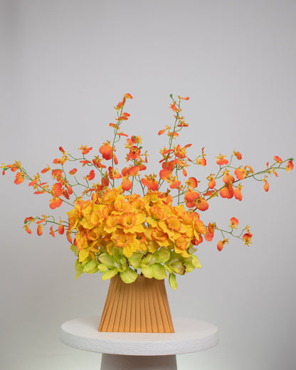 Vibrant artificial floral arrangement featuring orange orchids, yellow blossoms, and green cymbidiums in a pleated terracotta vase, styled on a white pedestal against a soft neutral background.