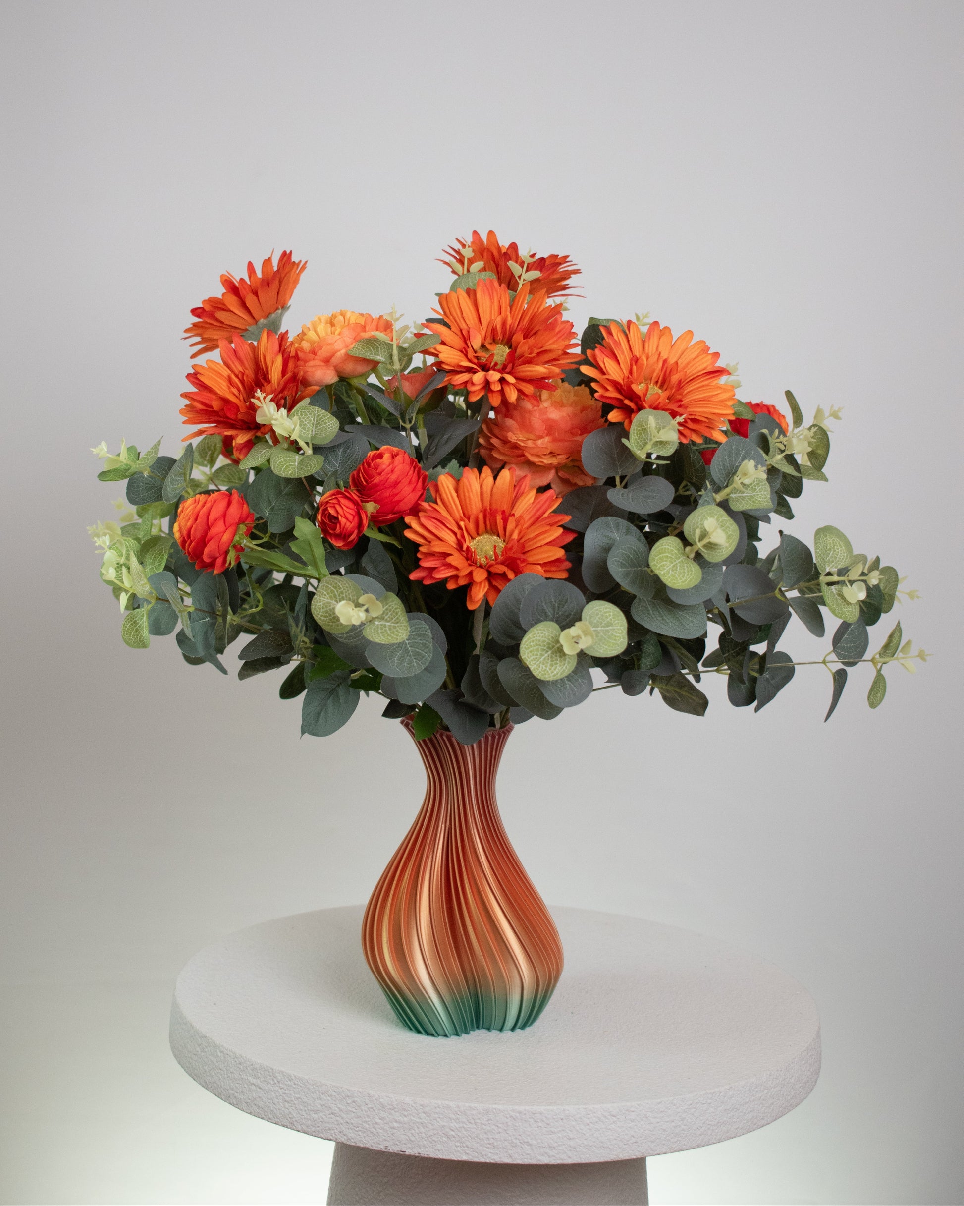 Artificial floral arrangement with orange gerberas and ranunculus surrounded by eucalyptus leaves, displayed in an ombre vase blending amber and green tones on a light background.