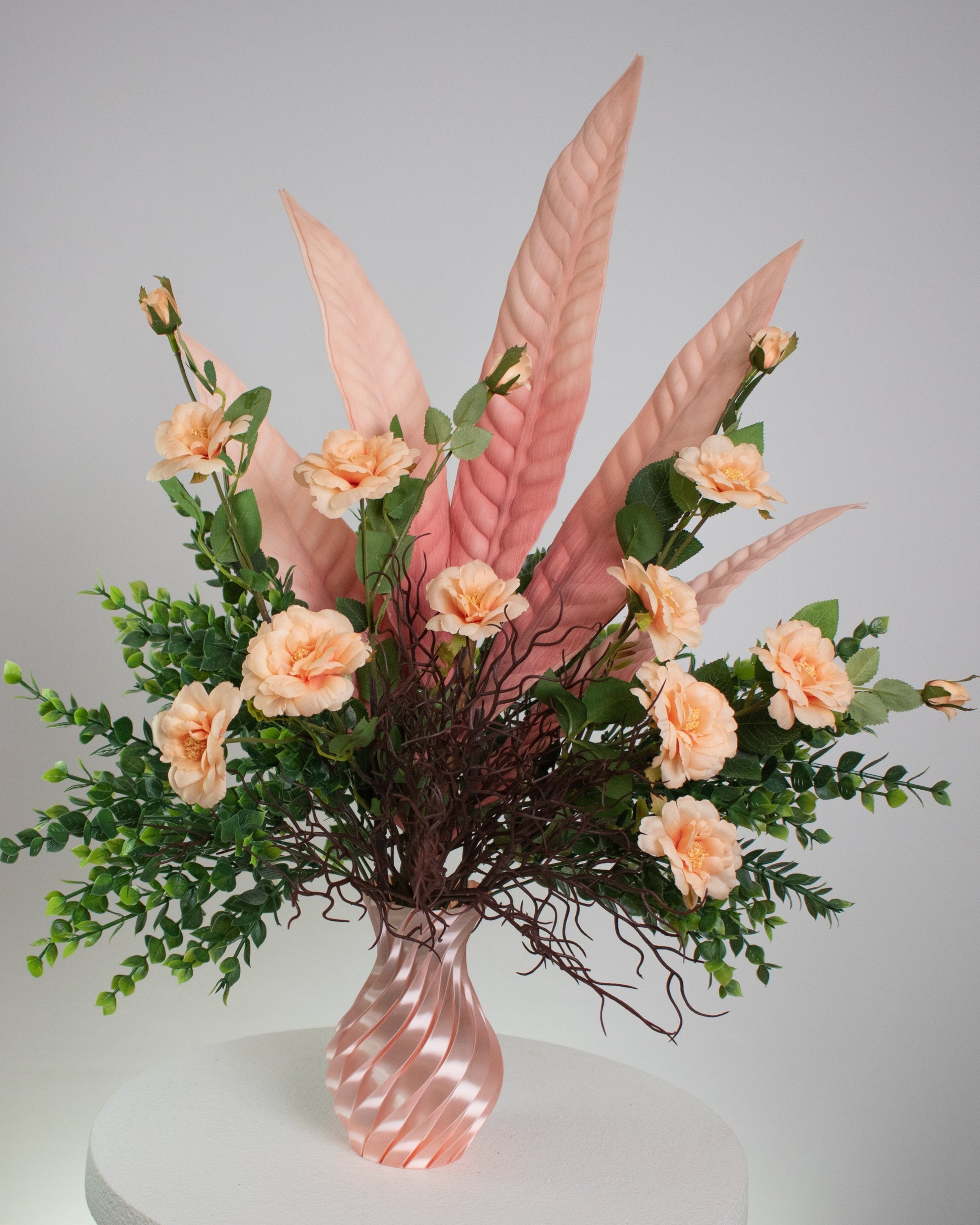 Artificial floral arrangement with peach roses, pink tropical leaves, and lush greenery in a twisted pink spiral vase, displayed on a white pedestal — perfect for elegant or modern interior spaces.