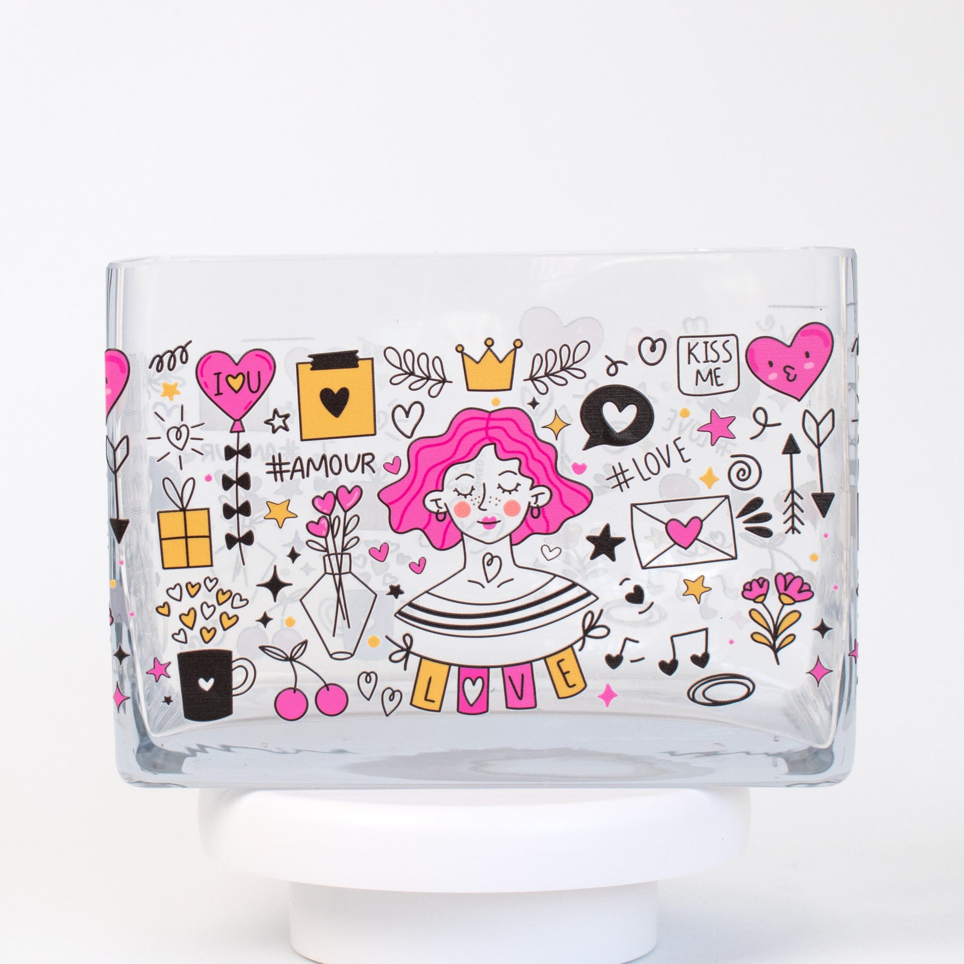 A clear rectangular glass vase featuring a colourful illustrated design of a pink-haired woman surrounded by hearts, flowers, love letters, and doodles in pink, gold, and black tones.