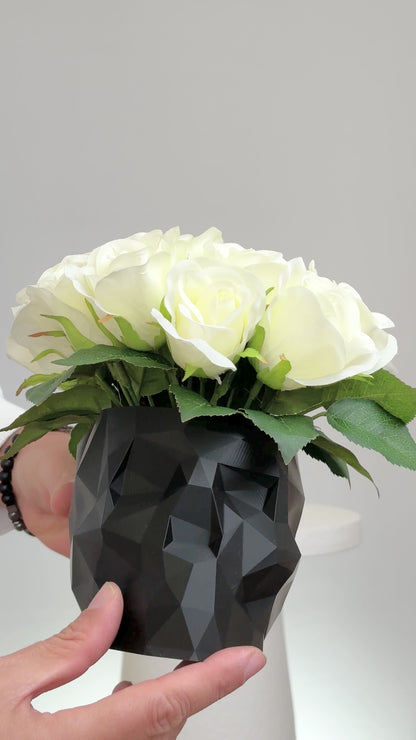 Artificial ivory rose arrangement displayed in a black geometric skull vase on a white pedestal against a neutral background.