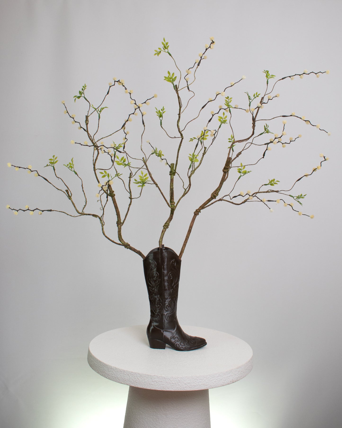 Rustic artificial floral arrangement with natural branches and light green leaves arranged in a dark brown cowboy boot-shaped vase, styled as a modern rustic décor accent.