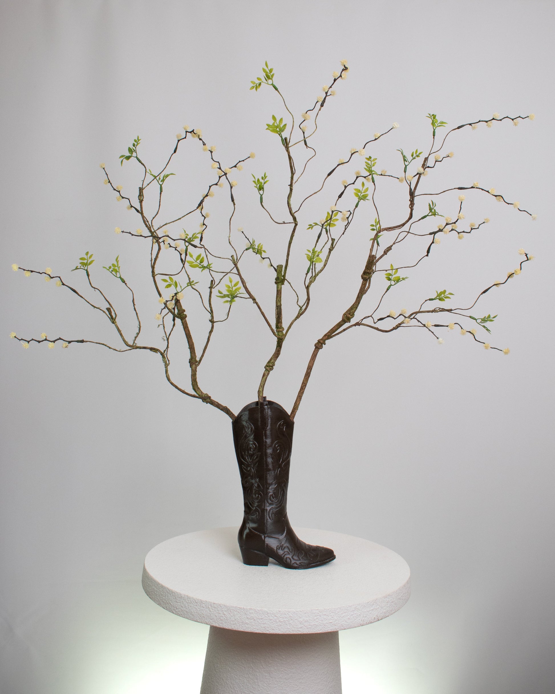 Rustic artificial floral arrangement with natural branches and light green leaves arranged in a dark brown cowboy boot-shaped vase, styled as a modern rustic décor accent.