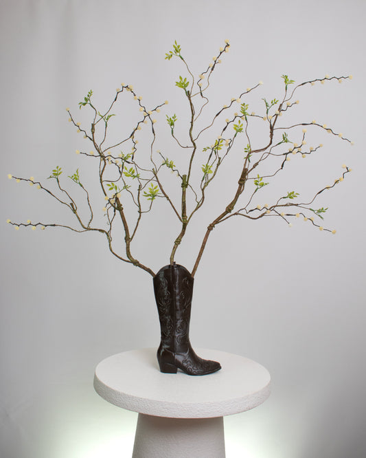 Rustic artificial floral arrangement with natural branches and light green leaves arranged in a dark brown cowboy boot-shaped vase, styled as a modern rustic décor accent.
