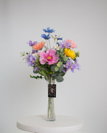 A vibrant artificial floral arrangement featuring pink cosmos, yellow and lavender blooms, peach gerberas, blue and white wildflowers, and green cherry blossom leaves, elegantly displayed in a clear glass vase with acrylic water, set against a neutral background.
