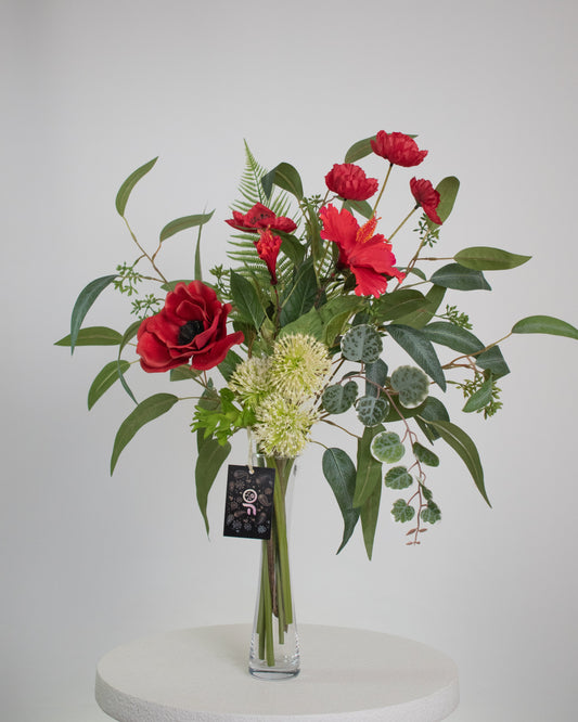 Artificial red floral arrangement with lush greenery and cream accents, displayed in a tall clear glass vase with acrylic water