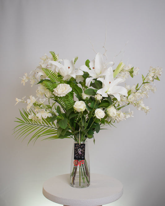 Elegant artificial floral arrangement featuring white lilies, roses, and orchids with lush green foliage in a clear glass vase — styled for a timeless and sophisticated look.