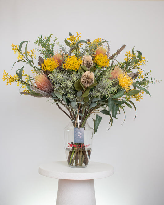 Artificial native flower arrangement with yellow pincushion proteas, banksia, and greenery, arranged in a clear glass vase with acrylic water