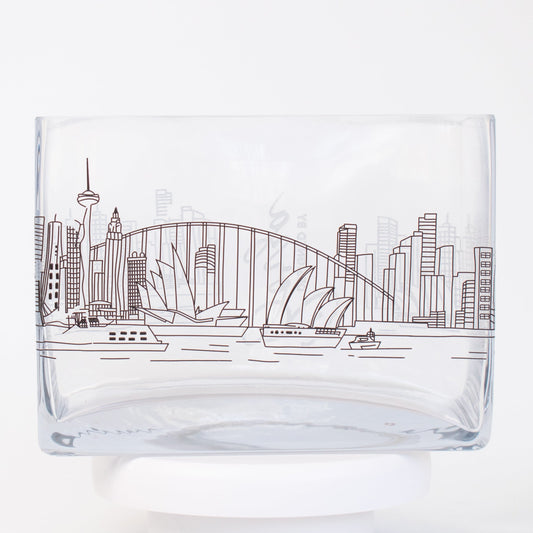 A clear rectangular glass vase featuring a fine-line illustration of the Sydney skyline, including the Opera House, Harbour Bridge, and city buildings.