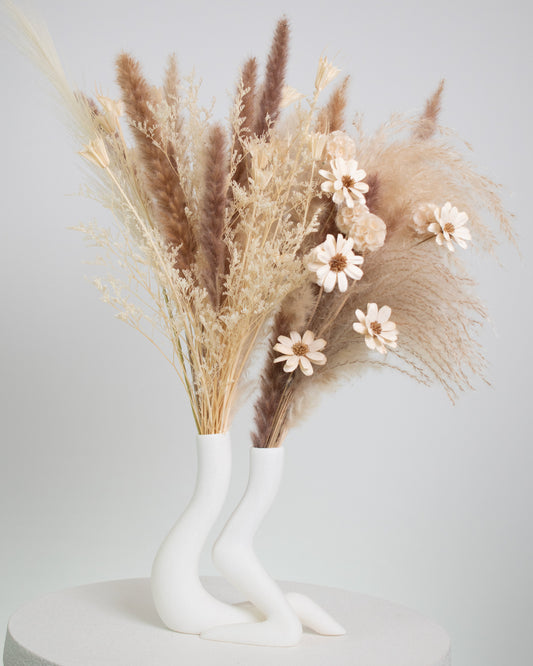 A modern dried floral arrangement featuring pampas grass, bunny tails, preserved foliage, and neutral-toned dried flowers, artistically displayed in a sculptural white ceramic double vase shaped like two flowing figures.