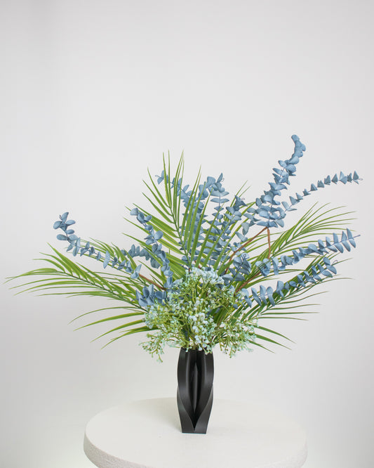 Artificial arrangement with blue eucalyptus stems, palm leaves, and delicate greenery in a sleek black sculpted vase, styled on a white pedestal — ideal for modern or minimalist interiors with cool or neutral tones.