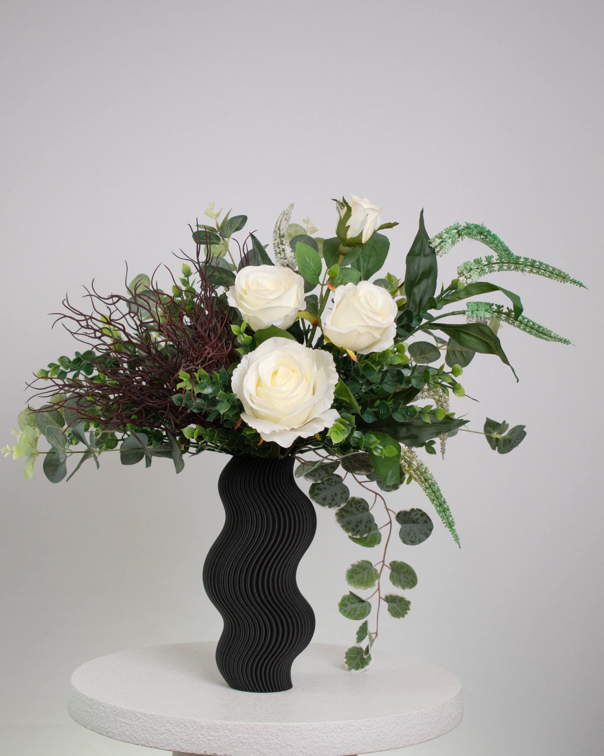 Artificial white rose arrangement with lush greenery in a sculptural matte black wavy vase, styled on a marble table — a modern and elegant decorative floral display ideal for minimalist interiors.