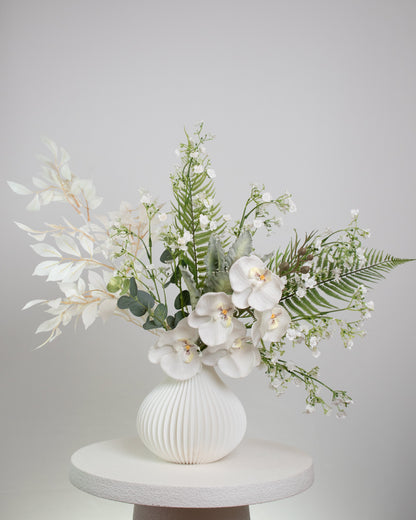 White orchid and foliage artificial flower display in sculpted white vase, minimalist interior décor.