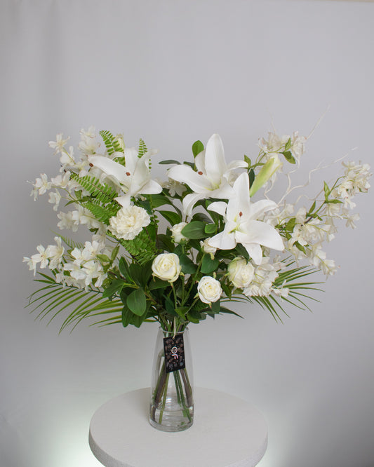 Elegant artificial floral arrangement featuring white lilies, roses, and orchids with lush green foliage in a clear glass vase — styled for a timeless and sophisticated look.